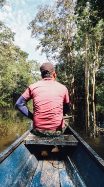 LETICIA, AMAZONAS, COLOMBIA - CRCA Aralık 2019: Güneşli bir günde Amazon Nehri 'nde yelken açan kırmızı gömlekli balıkçı teknenin tepesinde oturuyor..