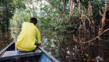LETICIA, AMAZONAS, COLOMBIA - CRCA Aralık 2019: Güneşli bir günde Amazon ormanlarında yelken açan motorlu teknenin üstündeki sarı tişörtlü çocuk.