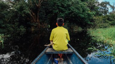 LETICIA, AMAZONAS, COLOMBIA - CRCA Aralık 2019: Güneşli bir günde Amazon ormanlarında yelken açan motorlu teknenin üstündeki sarı tişörtlü çocuk.