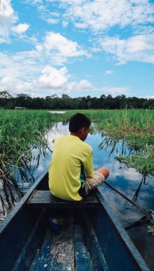 LETICIA, AMAZONAS, COLOMBIA - CRCA Aralık 2019: Güneşli bir günde Amazon 'da yeşil çimenler ve bitkilerle kaplı olarak göl boyunca yelken açan motorlu teknenin üzerindeki sarı tişörtlü çocuk.