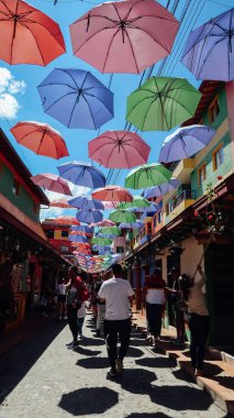 GUATAPE, COLOMBIA - CIRCA JANUARY 2020: İnsanlar renkli otantik Guatape şehrinde geleneksel eşsiz binalar ve şemsiyelerle kaldırım taşı caddelerinde yürüyor.