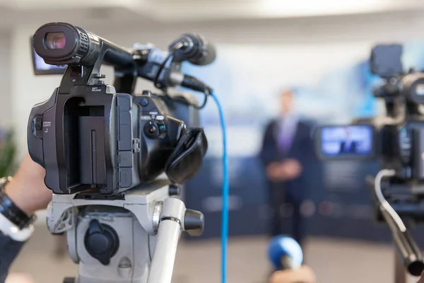 Video camera in focus, blurred spokesman in background — Stock Photo, Image