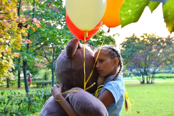 Грустная девочка обнимает большого плюшевого мишку и воздушные шарики в парке. Школьница несчастна в парке с большой игрушкой и мячами . — стоковое фото