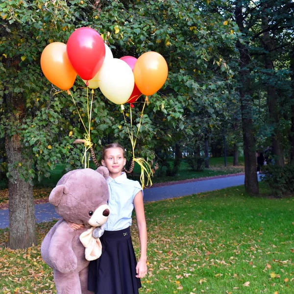 Грустная девочка обнимает большого плюшевого мишку и воздушные шарики в парке. Школьница несчастна в парке с большой игрушкой и мячами . — стоковое фото