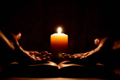 Náboženská modlitba se svíčkou v naprosté tmě. Požádej Boha o pomoc. Modlí se v Bibli. Dlaně na staré knize na černém pozadí. Kniha, svíčky a ruce. Žena se modlí ve tmě. Čtení modliteb v noci.