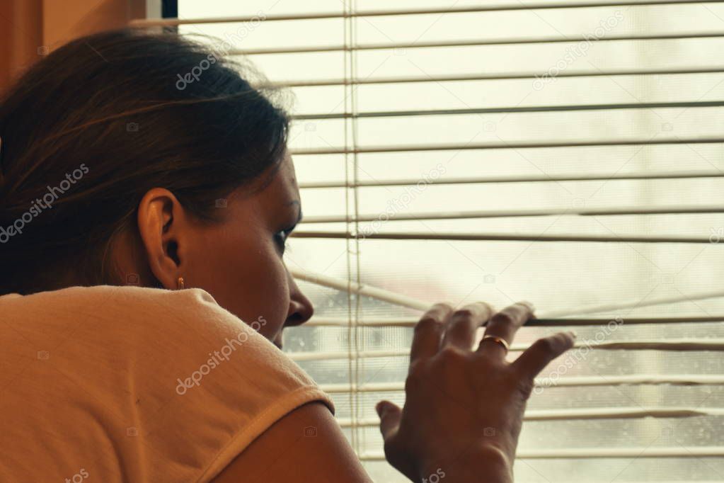 A woman peeps out the window through the blinds.