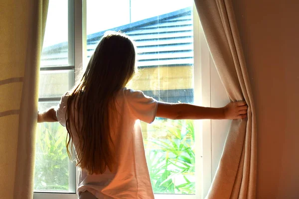 Dziewczyna otworzyła zasłony i wyglądała przez okno. Rano w oknie świeci słońce.. — Zdjęcie stockowe