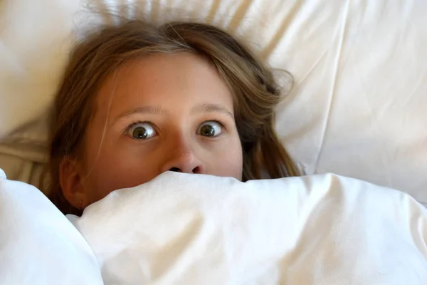 Meisje kind kijkt uit van onder de dekens. — Stockfoto