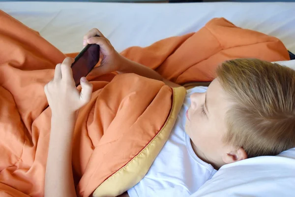 Tiener jongen speelt aan de telefoon in het bed. Mobiele verslaving van kinderen — Stockfoto