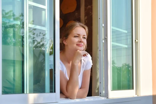 Piękna kobieta przy oknie. Szczęśliwa dziewczyna wygląda przez otwarte okno — Zdjęcie stockowe