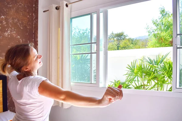 Een meisje wordt wakker bij het open raam. En ochtendzonlicht. een nieuwe dag om te glimlachen. Genieten van het leven. — Stockfoto