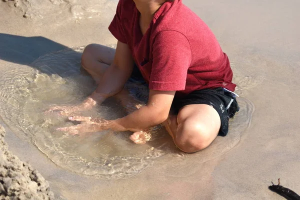 Ein Junge spielt in einer Pfütze am Strand. Das Kind sitzt in einer Pfütze im Sand. fröhlicher lustiger Sommer. — Stockfoto