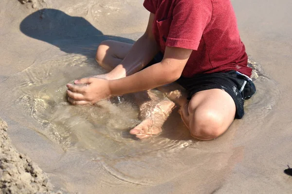 Un niño juega en un charco en la playa. El niño se sienta en un charco en la arena. Feliz verano divertido . — Foto de Stock