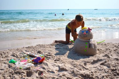 Child boy makes a Sandman on the beach. The child builds a snowman on the beach. New year on the tropical ocean. clipart