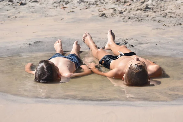 Два маленьких мальчика сидят в луже. Дети играют в водяной луже. Два брата на пляже в песке. Счастливого беззаботного лета в море. — стоковое фото