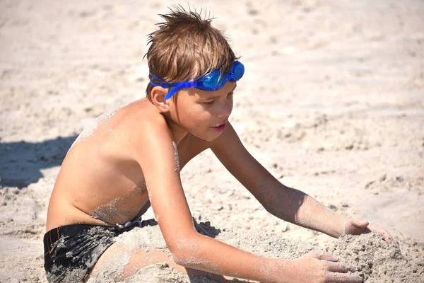 O rapazinho na praia brinca. A criança nos copos de água no mar . — Fotografia de Stock