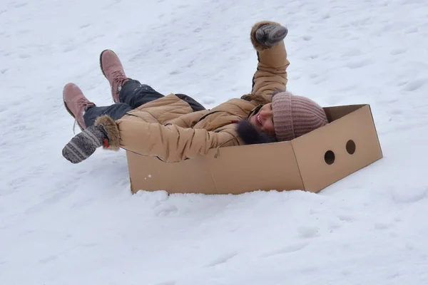 Ragazza in una scatola di cartone sulla collina in inverno. Bambino per divertirsi in inverno. L'inverno è un gioco divertente. Vacanze sportive a Natale . — Foto Stock