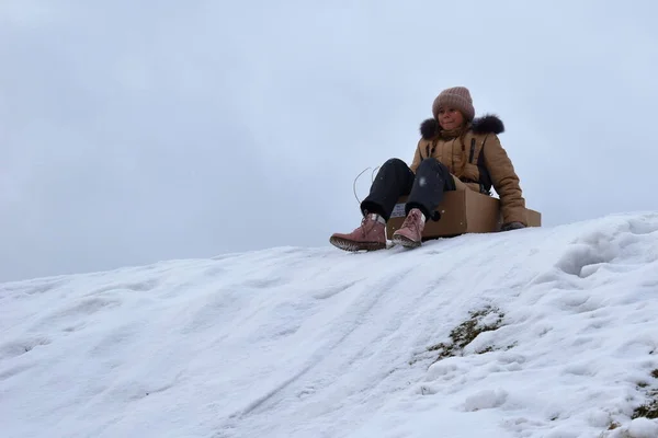 Ragazza in una scatola di cartone sulla collina in inverno. Bambino per divertirsi in inverno. L'inverno è un gioco divertente. Vacanze sportive a Natale . — Foto Stock