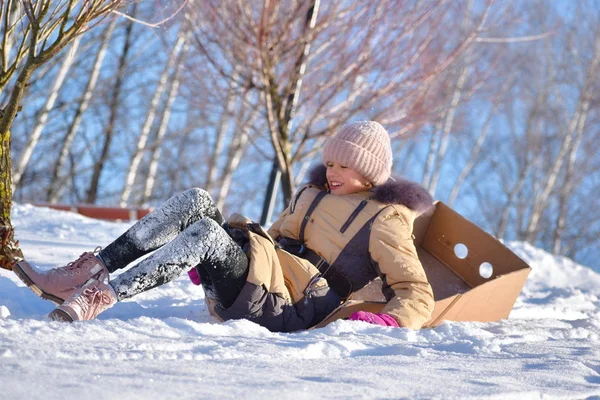 Menina em uma caixa de papelão. Deslize de inverno com a criança. Andar na neve. montando na caixa — Fotografia de Stock