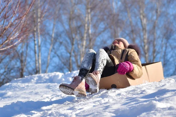 Una chica en una caja de cartón. Tobogán de invierno con el niño. Cabalgando en la nieve. montar en la caja —  Fotos de Stock