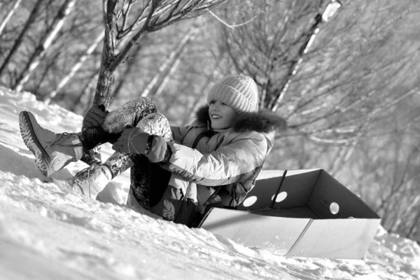 Foto monocromática. Una chica en una caja de cartón. Tobogán de invierno con el niño. Cabalgando en la nieve. montar en la caja —  Fotos de Stock