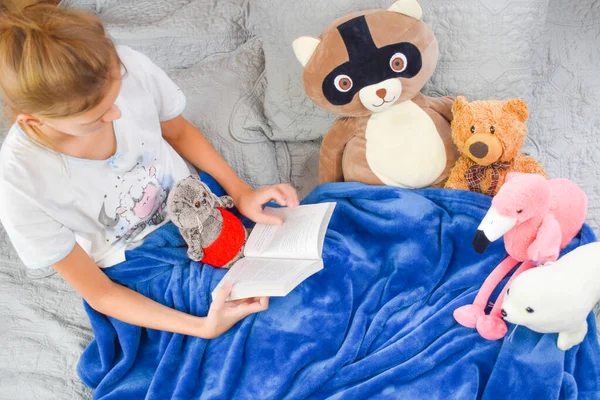 Thuisonderwijs. Schoolmeisje leest leerboek op het bed. Baby meisje het lezen van een verhaal aan speelgoed — Stockfoto