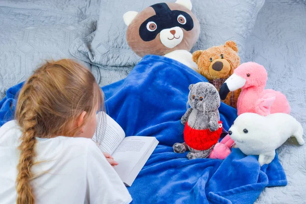 Sekolah di rumah. Gadis sekolah membaca buku teks di tempat tidur. Bayi perempuan membaca cerita untuk mainan — Stok Foto