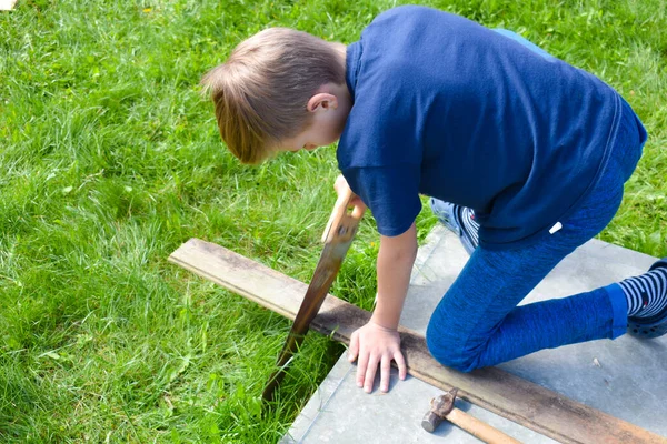 Мальчик пилит дрова. Мальчик работает в саду. Детская мастерская снаружи — стоковое фото