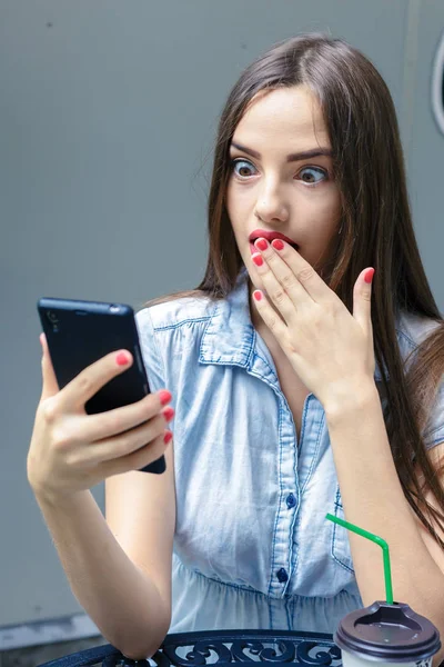 Mujer joven sorprendida mirando teléfono inteligente en la cafetería al aire libre — Foto de Stock