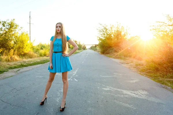 Mooi blonde meisje in blauwe jurk permanent op een weg bij zonsondergang — Stockfoto