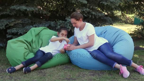 Mãe e filha colocando moedas em um banco rosa piggy sentado em almofadas no parque em um dia de verão . — Vídeo de Stock