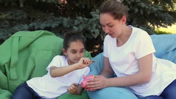 Mother and daughter putting coins into a pink piggy bank sitting on cushions in the park on a summer day. — Stock Video