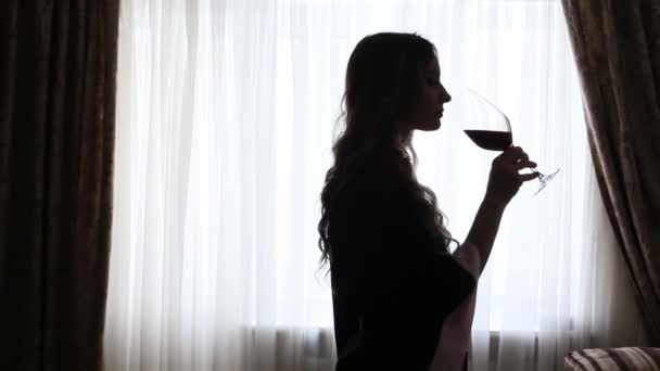 Muchacha joven vestida con una bata bebe un vino de vino grande, silueta en una luz de fondo sobre un fondo de ventana — Vídeos de Stock