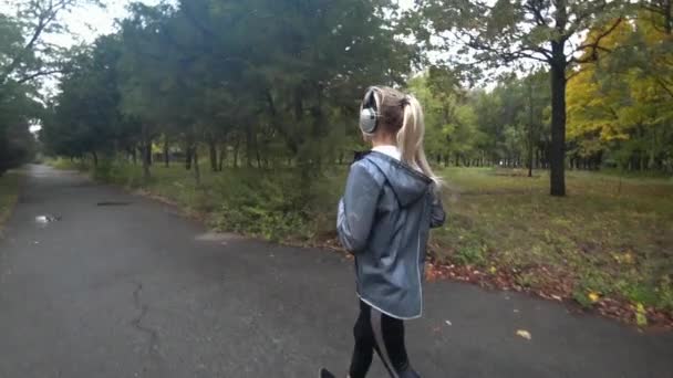 Hermosa joven rubia con el pelo largo se ejecuta en el parque en el día lluvioso, escuchar música en los auriculares, hacer deportes, un estilo de vida saludable.. Movimiento lento . — Vídeo de stock