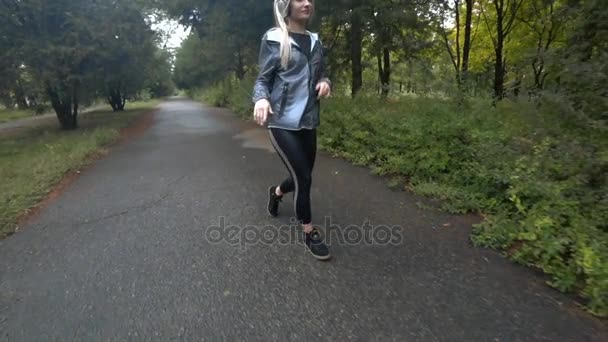 Hermosa joven rubia con el pelo largo caminando en el parque, escuchar música en un día de otoño lluvioso.. Movimiento lento . — Vídeos de Stock