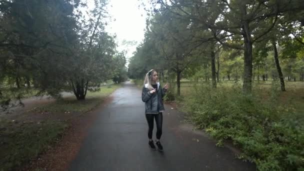 Schöne junge blonde Mädchen mit langen Haaren, die im Park spazieren gehen, Musik hören an einem regnerischen Herbsttag.. Zeitlupe. — Stockvideo