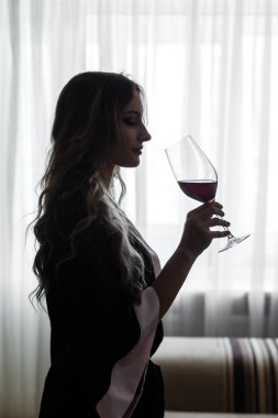Bir elbise giymiş şehvetli genç kadın büyük bardağı, siluet bir pencere arka planda bir arka ışık içinde gelen bir kırmızı şarap içer