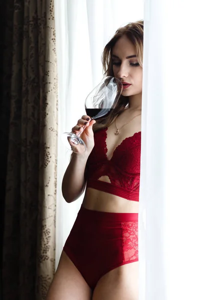 Чуттєва молода жінка, одягнена в червону білизну, п'є вино вікно . — стокове фото