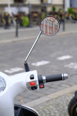 Kentsel beyaz scooter şehrin turistik merkezindeki kaldırım taşı yolunda park halinde. Kulplu kontroller, aynada eski bir evin yansıması..