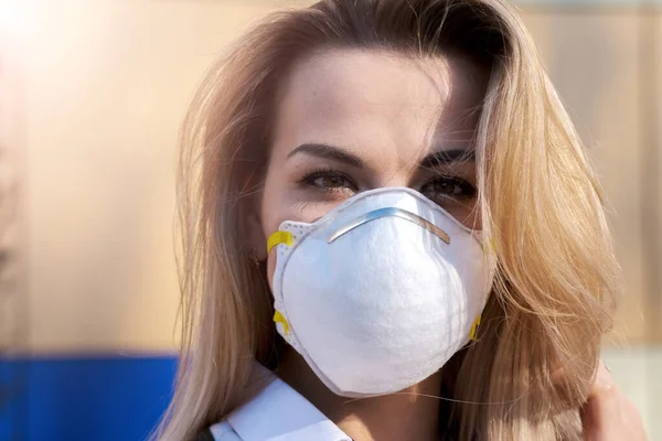 Genç, güzel, sarışın, beyaz bir kadın koruyucu maske takmış, kameraya bakıyor..