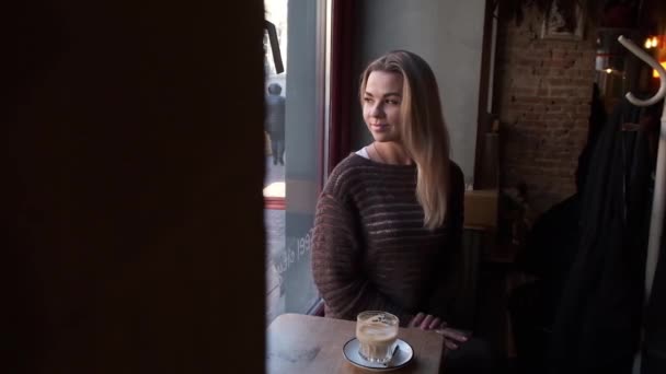若いですかわいいですブロンドの髪を持つ女性カフェの窓の近くに座ります コーヒーを飲みます — ストック動画