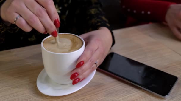 小女孩把咖啡和白杯中的泡沫混在一起 坐在一个有智能手机的桌子旁的咖啡店里 有红色指甲特写的手 — 图库视频影像