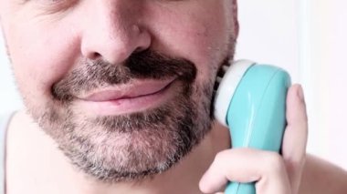 40 yaşında orta yaşlı, sakallı bir adamın yüz masajı yapmak için taşınabilir elektrikli masaj aleti kullanması. Sağlıklı cilt bakımı, erkek güzellik konsepti.