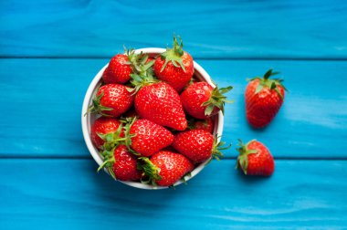 Mavi ahşap arka planda beyaz bir kasede olgun çilekler. Sağlıklı taze meyveler. Üst görünüm