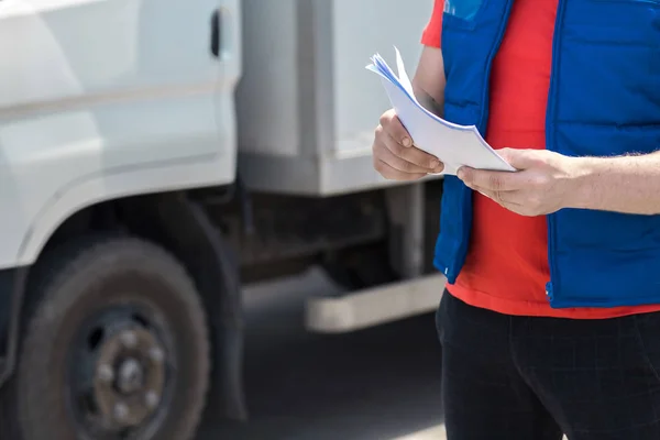 Courier driver in uniform making notes in document and delivery white truck behinde him