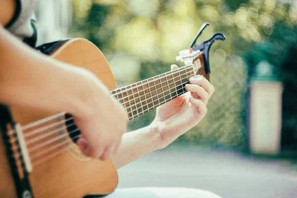 Close up guitar neck with capo in the park. Young man sitting on the bench in the park playing the guitar. Young attractive man enjoys live music in last sunny days autumn holiday. Retro lens used. — Stock Photo, Image