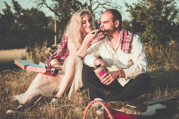 Pareja romántica abrazándose mientras hace un picnic bajo la luz del atardecer. Atractiva joven mujer alimentando a su hombre con una pizza. Están sentados juntos en la hierba al aire libre . — Foto de Stock