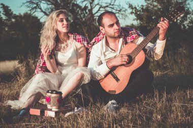 Romantik çift gün batımında piknik yaparken sarılıyor. Gitarlı bir adam ve beyaz elbiseli çekici genç bir kadın açık havada çimlerin üzerinde birlikte oturuyorlar..