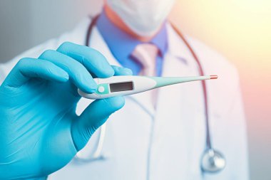 Gazlı bez maskeli bir doktor ve elinde mavi kauçuk eldiveniyle termometre tutuyor.