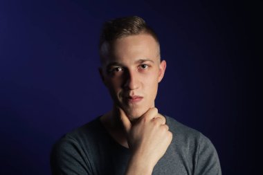 Koyu mor arka planda gri tişörtlü, beyaz, genç bir adam çenesine dokunuyor ve bir şeyler düşünüyor.